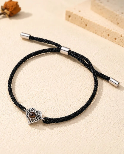 Armband mit Bild im Stein: silberfarbenes Herz aus Engelsflügeln an schwarzer Kordel auf hellem Gips
