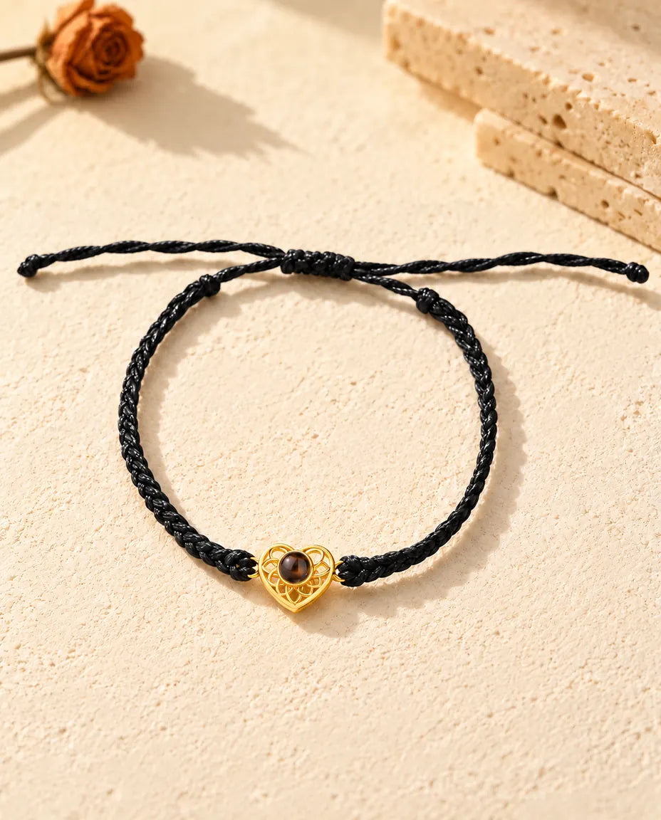 Draufsicht: Armband mit goldfarbenem Herz an schmalem schwarzem Flechtband, mit Schiebeknoten und Bandenden