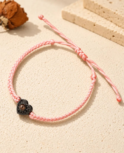 Armband mit schmalem rosa Flechtband, schwarzem Herz und braunem Bildstein auf hellem Gips