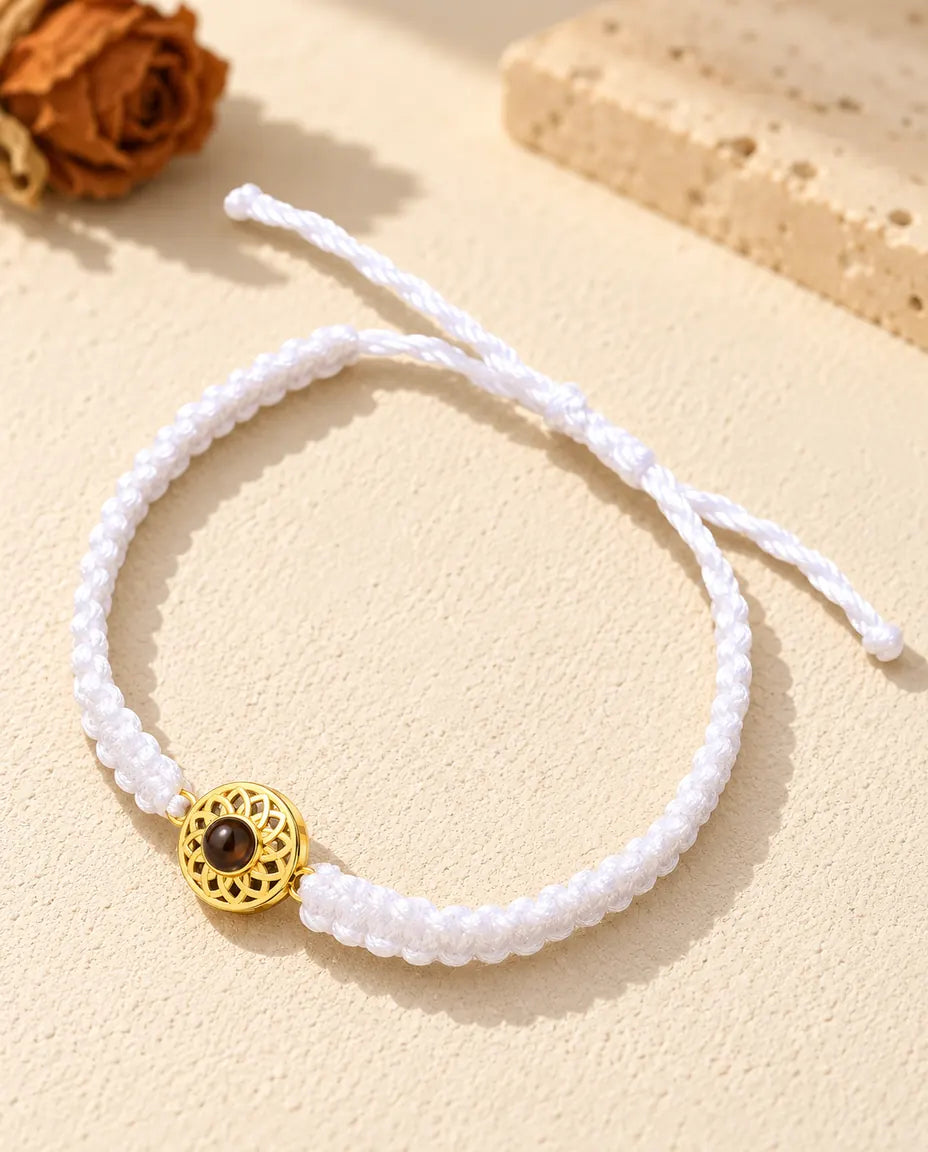 Armband mit weißem Flechtband, goldfarbenem Rund-Charm und Schiebeknoten auf hellem Gips