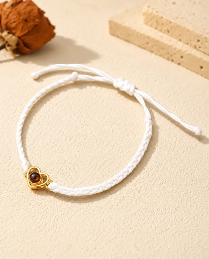 Armband mit weißem Flechtband, goldfarbenem offenem Herz und Schiebeknoten auf hellem Gips